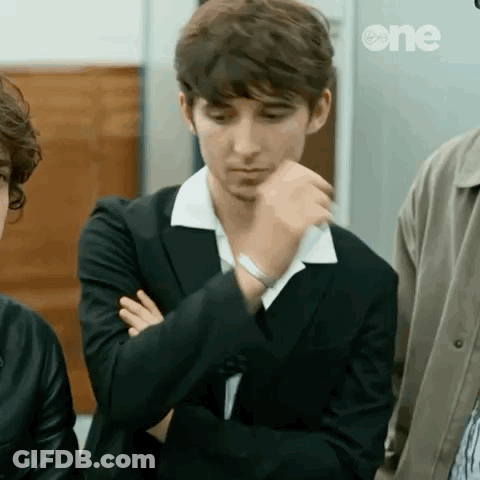 A Young Man In A Suit And White Collar Is Sitting With His Arms Crossed And His Hand On His Chin . GIF