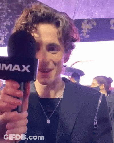 A Young Man In A Suit Is Holding A Microphone In His Hand And Smiling . GIF