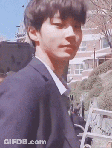 A Young Man In A Suit Is Standing In Front Of A Building Looking At The Camera . GIF