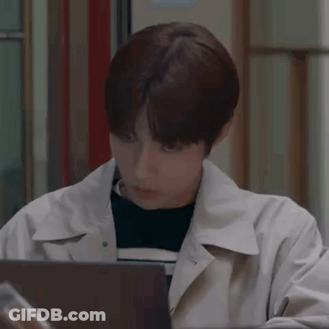 A Young Man In A Trench Coat Is Sitting In Front Of A Laptop . GIF