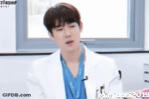 A Young Man In A White Coat And Blue Scrub Is Sitting In Front Of A Window . GIF