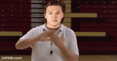 A Young Man In A White Shirt Is Making A Gesture With His Hands In Front Of A Basketball Court . GIF