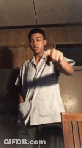 A Young Man In A White Shirt Is Standing In Front Of A Chair GIF