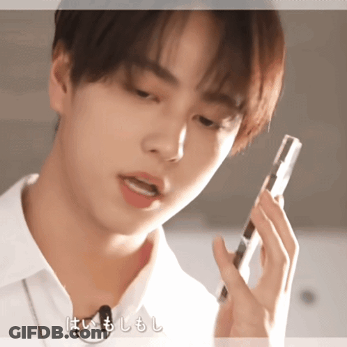 A Young Man In A White Shirt Is Talking On A Cell Phone With Chinese Writing Behind Him GIF