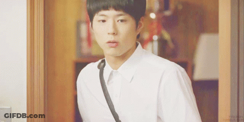 A Young Man In A White Shirt With A Black Strap Around His Neck Is Standing In Front Of A Door GIF