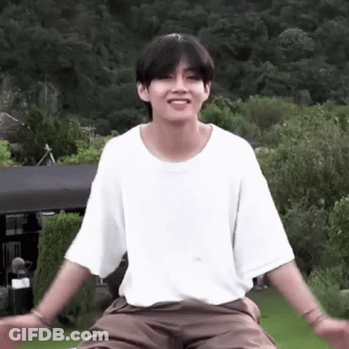 A Young Man In A White T-Shirt And Brown Shorts Is Sitting On A Lawn With His Arms Outstretched . GIF