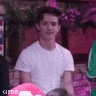 A Young Man In A White T-Shirt Is Sitting In Front Of A Pink Heart Pillow . GIF