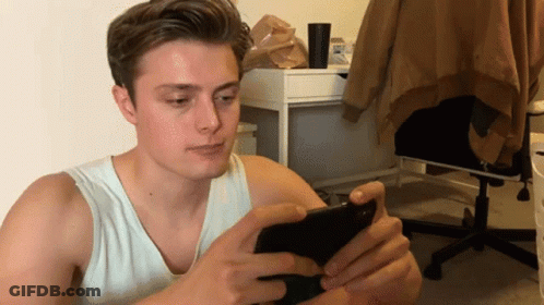 A Young Man In A White Tank Top Is Looking At His Phone GIF