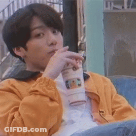 A Young Man In A Yellow Jacket Is Sitting On A Bench Drinking From A Cup . GIF