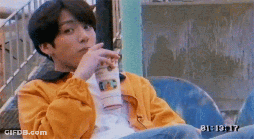 A Young Man In A Yellow Jacket Is Sitting On A Chair Drinking A Cup Of Coffee . GIF