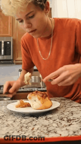 A Young Man In An Orange Shirt Is Eating A Cinnamon Roll GIF
