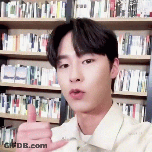 A Young Man Is Giving A Thumbs Up Sign In Front Of A Bookshelf . GIF
