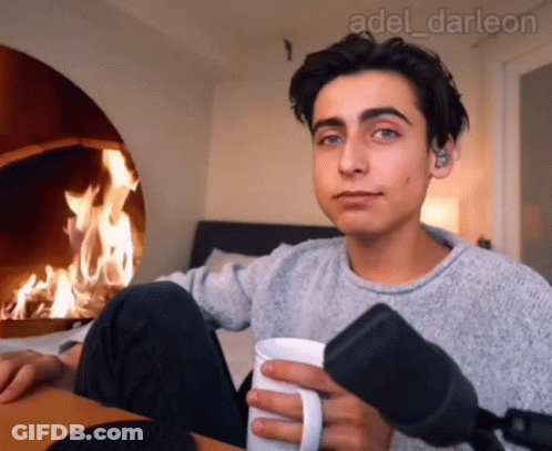 A Young Man Is Holding A Cup Of Coffee In Front Of A Fireplace GIF