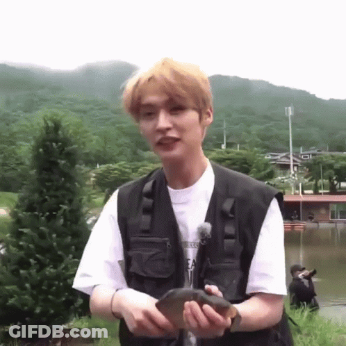 A Young Man Is Holding A Fish In His Hands While Standing In Front Of A Lake . GIF