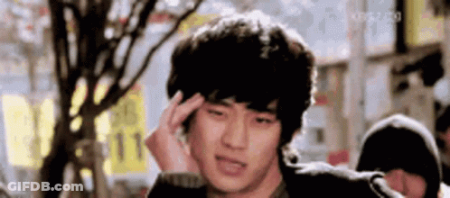 A Young Man Is Holding His Head In Pain While Standing In Front Of A Tree . GIF
