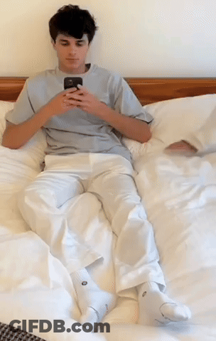 A Young Man Is Laying On A Bed Looking At His Cell Phone GIF