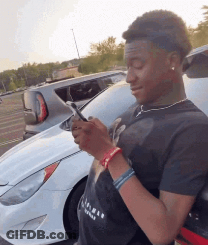 A Young Man Is Looking At His Phone In Front Of A Car GIF