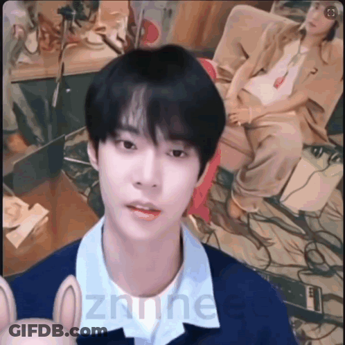 A Young Man Is Looking At The Camera While A Woman Sits Behind Him On A Couch . GIF