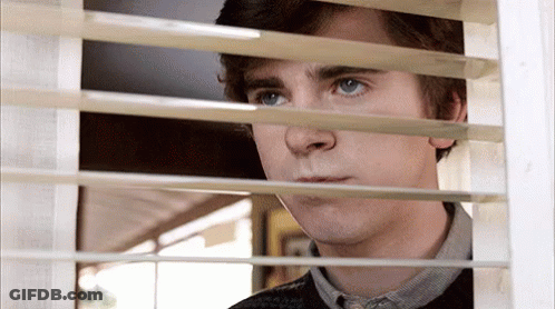 A Young Man Is Looking Out Of A Window With Blinds . GIF