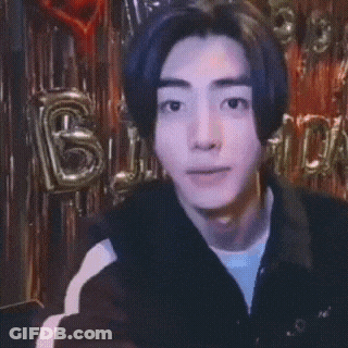 A Young Man Is Pointing At The Camera With Balloons Behind Him That Say Birthday GIF