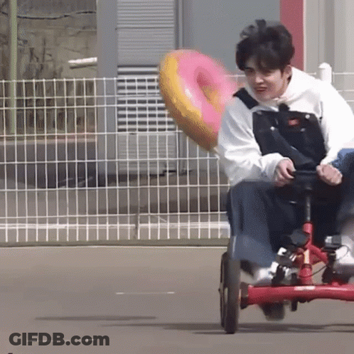 A Young Man Is Riding A Tricycle With A Pink Donut In The Back . GIF