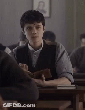 A Young Man Is Sitting At A Desk In A Classroom Holding A Book GIF