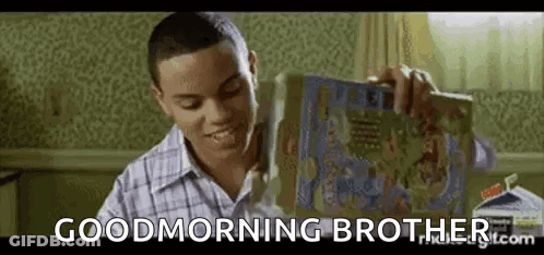 A Young Man Is Sitting At A Table Holding A Box Of Cereal And Smiling . GIF