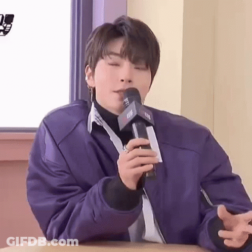 A Young Man Is Sitting At A Table Holding A Microphone In His Hand . GIF