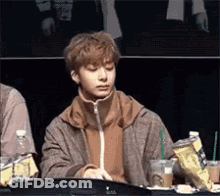A Young Man Is Sitting At A Table With A Drink In His Hand And A Bag Of Chips . GIF