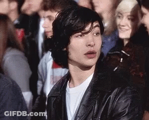 A Young Man Is Sitting In A Crowd Of People Wearing A Leather Jacket . GIF
