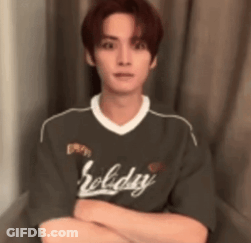 A Young Man Is Sitting In Front Of A Window With His Arms Crossed And Wearing A Holiday Shirt . GIF