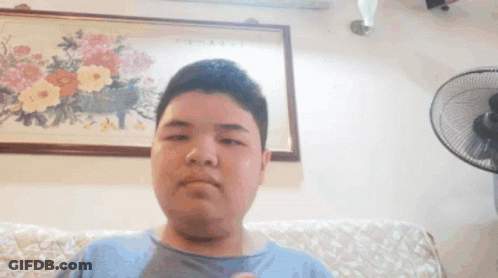 A Young Man Is Sitting On A Couch In Front Of A Fan And A Painting Of Flowers On The Wall . GIF