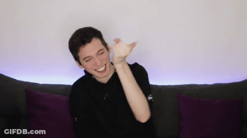 A Young Man Is Sitting On A Couch With Purple Pillows And Smiling GIF