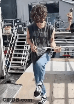 A Young Man Is Sitting On A Wooden Platform Holding A Drum Stick . GIF