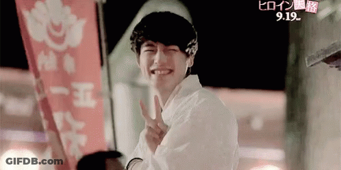 A Young Man Is Smiling And Giving A Peace Sign While Standing In Front Of A Red Banner . GIF