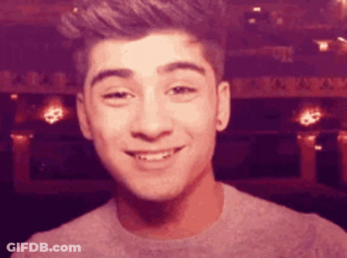 A Young Man Is Smiling And Looking At The Camera While Wearing A Gray Sweater . GIF