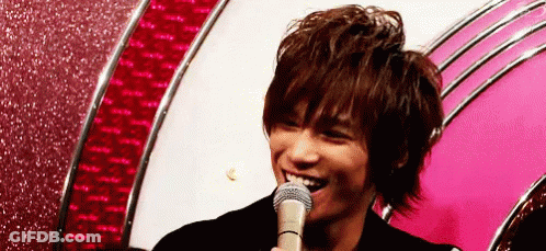 A Young Man Is Smiling While Holding A Microphone In Front Of A Pink Wall . GIF
