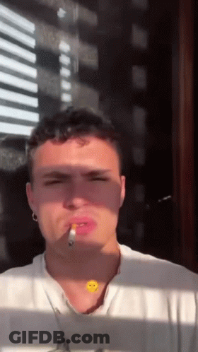 A Young Man Is Smoking A Cigarette In Front Of A Window While Wearing A Smiley Face Necklace . GIF