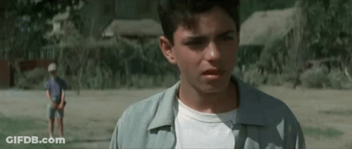 A Young Man Is Standing In A Field Looking At The Camera While Another Young Man Stands In The Background . GIF