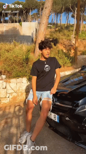 A Young Man Is Standing In Front Of A Black Car . GIF