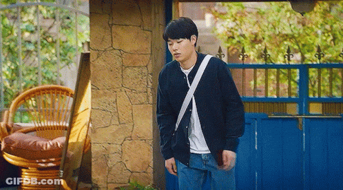 A Young Man Is Standing In Front Of A Blue Fence And A Wicker Chair . GIF