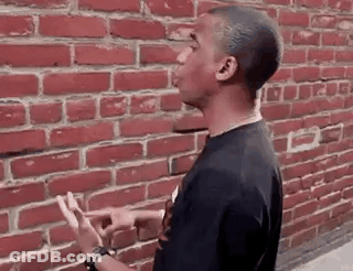 A Young Man Is Standing In Front Of A Red Brick Wall And Pointing At It . GIF