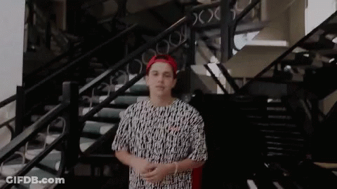 A Young Man Is Standing In Front Of A Set Of Stairs Wearing A Red Hat And A Black And White Shirt . GIF
