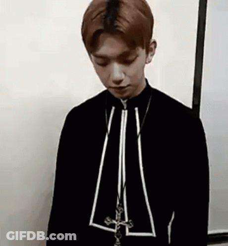 A Young Man Is Standing In Front Of A White Wall Wearing A Black Jacket And A Necklace . GIF