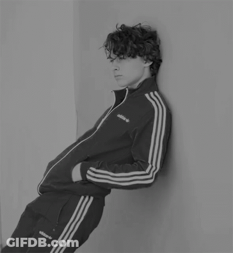 A Young Man Leaning Against A Wall Wearing An Adidas Tracksuit GIF