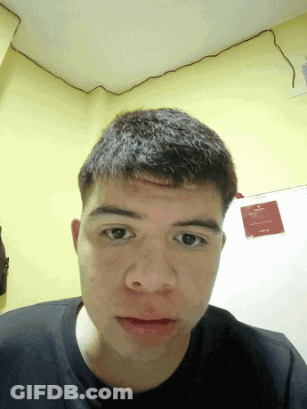A Young Man Looks At The Camera In Front Of A Yellow Wall With A Sign That Says " No Smoking " On It GIF