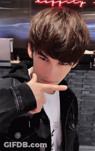 A Young Man Making A Peace Sign With His Finger In Front Of A Neon Sign That Says Happy GIF
