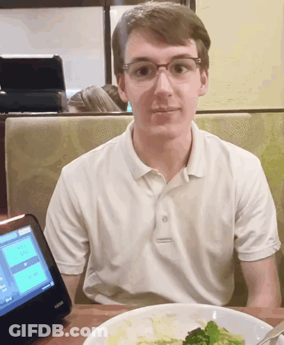 A Young Man Sits At A Table In Front Of A Kiosk That Shows A Menu GIF