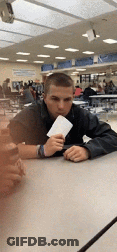 A Young Man Sitting At A Table In A Cafeteria Holding A Piece Of Paper GIF