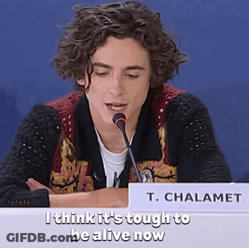 A Young Man Sitting At A Table With A Microphone And A Sign That Says T. Chalamet On It GIF